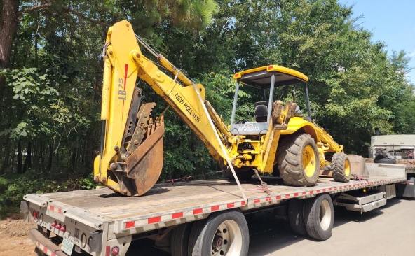 New Holland LB75 backhoe loader being transported on a flatbed trailer.