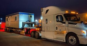 A white truck is transporting a white and blue office trailer